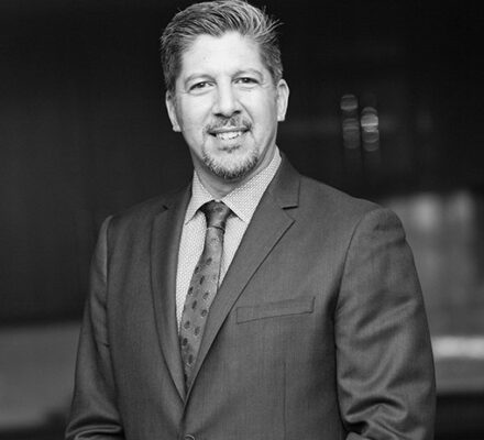 A man in a suit and tie stands facing the camera, smiling, with a blurred indoor background resembling an upscale appliance showroom. The image is in black and white.