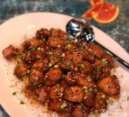 A plate of glazed chicken pieces garnished with chopped green onions and sesame seeds, served over white rice, with a black spoon on the side. Orange slices are in the background, evoking flavors found in a luxury kitchen appliance showroom.