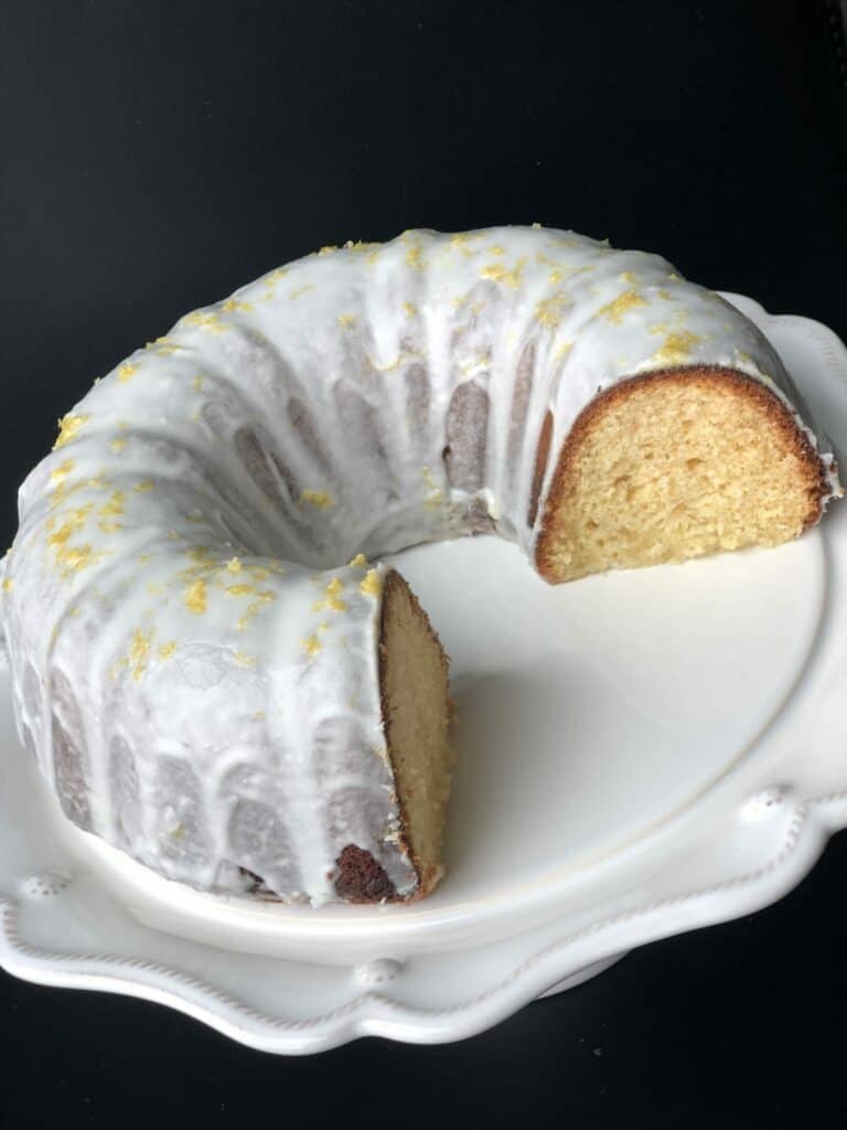 A lemon-glazed bundt cake with a slice removed, displayed on a white decorative plate against a dark background, evokes the elegance of a luxury kitchen appliance collection in a modern kitchen showroom.