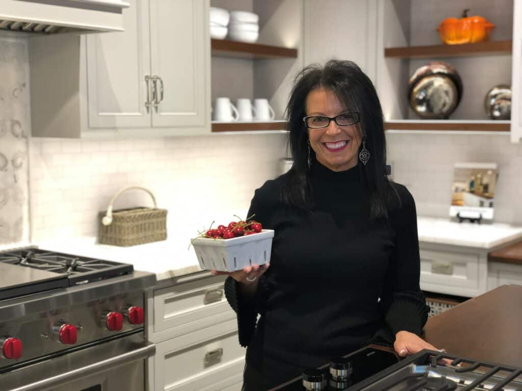 Woman in a black top holding a bowl of cherries stands in a modern kitchen with white cabinets and a Wolf luxury kitchen appliance.