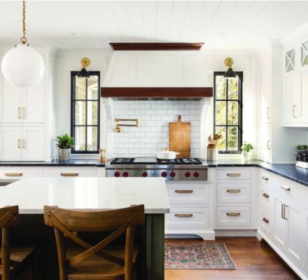 Modern kitchen with white cabinets, black countertops, stainless steel appliances including a Sub Zero appliance, a central island with wooden stools, brass fixtures, and two windows flanking a tiled backsplash.