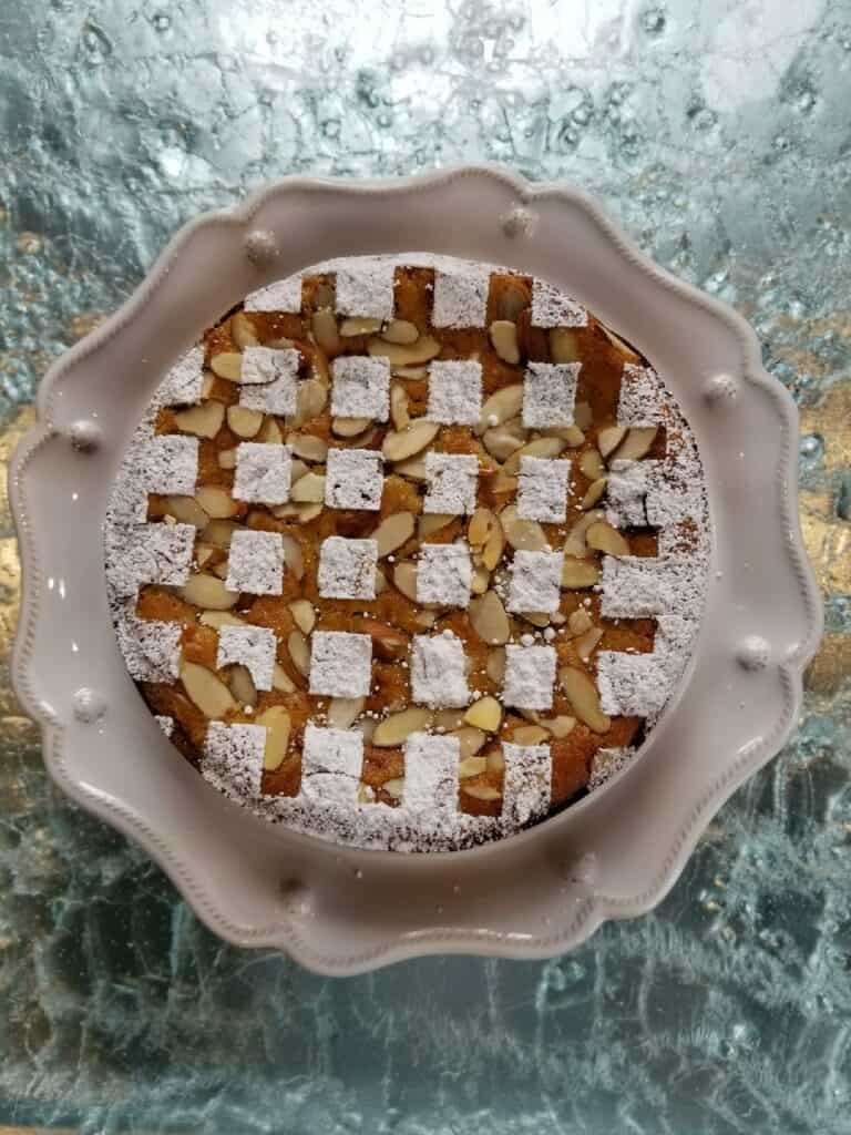 A round cake with a powdered sugar checkerboard pattern and sliced almonds on top, served on a white scalloped plate over a textured glass surface—perfectly baked using a luxury kitchen appliance.