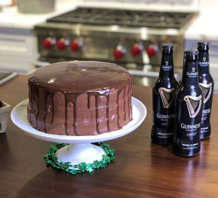A chocolate cake with glossy ganache sits on a white cake stand, beside four bottles of Guinness beer on a kitchen showroom countertop.