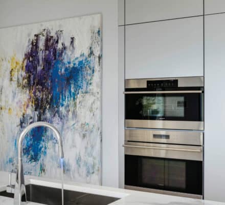 Modern kitchen with a marble countertop, built-in Wolf appliance ovens, a tall faucet, and an abstract painting with blue, purple, and yellow accents on the wall—perfect inspiration for any luxury kitchen appliance showroom.