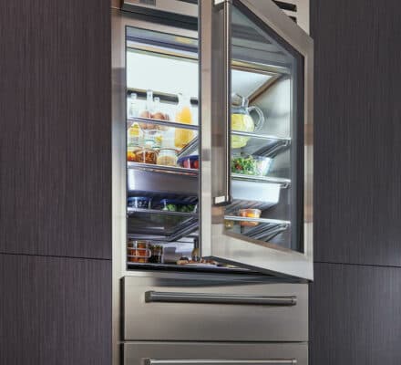 A stainless steel refrigerator with the door open, showing shelves stocked with food and drinks. This luxury kitchen appliance is built into dark wood cabinetry, creating an elegant display fit for an appliance showroom.