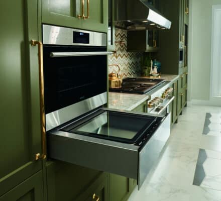 A modern kitchen showroom with green cabinets, a Wolf appliance wall oven open, and a gas cooktop, overlooking a window with a pool view.