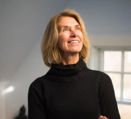 A woman with shoulder-length blonde hair wearing a black turtleneck sweater stands indoors, smiling and looking upward near a window in a modern kitchen showroom.