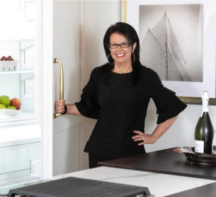A woman in black glasses and a black top stands in a modern kitchen showroom, holding open a refrigerator door. Fruit is visible inside, and a bottle of wine with glasses sits on the counter.