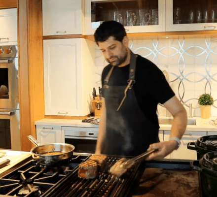 A man wearing an apron is grilling meat on a stovetop grill in a modern kitchen showroom with white cabinets and sleek Sub Zero appliances.