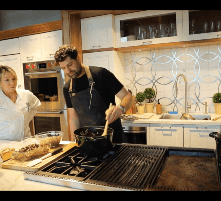 Two people stand in a modern kitchen showroom; one stirs food in a pot on the stove while the other looks on, with various ingredients and utensils displayed on the counter.
