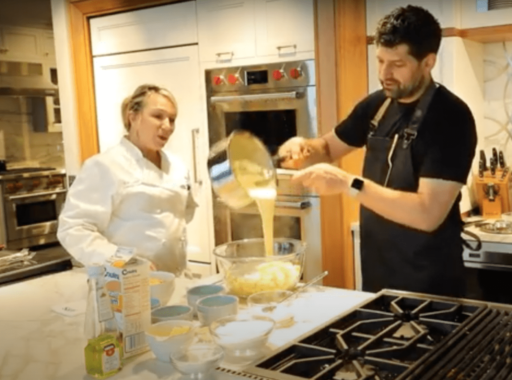 Two people in a kitchen showroom, one wearing a chef's coat, the other pouring batter from a pot into a mixing bowl surrounded by ingredients and a sleek Sub Zero appliance on the counter.