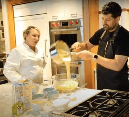 Two people in a kitchen showroom, one wearing a chef's coat, the other pouring batter from a pot into a mixing bowl surrounded by ingredients and a sleek Sub Zero appliance on the counter.