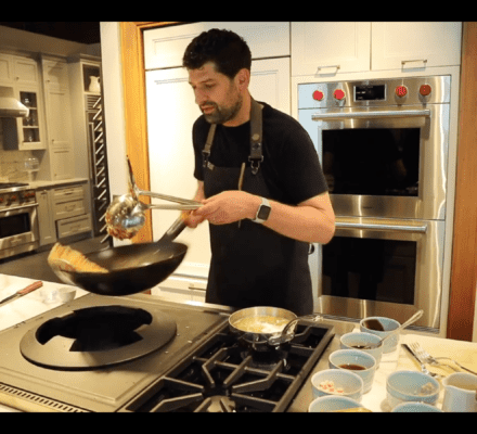 A man in a kitchen holds a wok and metal tongs over a wolf appliance stove, preparing food with several small bowls of ingredients arranged on the counter.