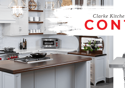 Modern white kitchen with stainless appliances, a Sub-Zero appliance, and wood countertops features a stove island. Promo reads "Clarke Kitchen Envy Design Contest" with a grill pictured on the right, highlighting our premier kitchen showroom.