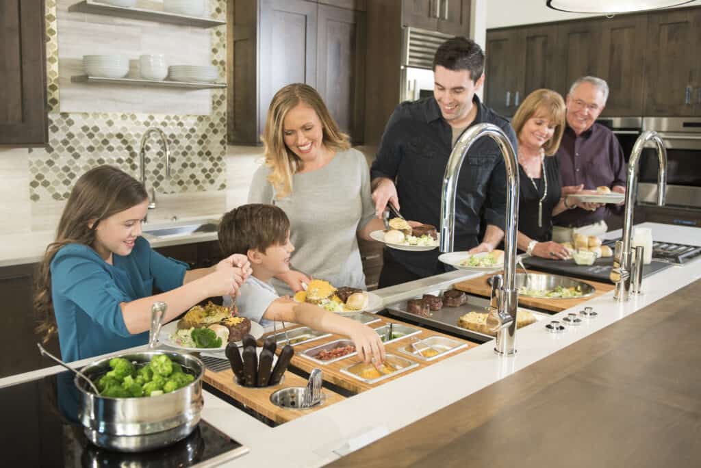 Six people serve themselves food from a buffet-style counter in a modern kitchen showroom, smiling and interacting as they prepare their plates beside sleek Wolf appliances.