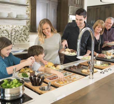 Six people serve themselves food from a buffet-style counter in a modern kitchen showroom, smiling and interacting as they prepare their plates beside sleek Wolf appliances.