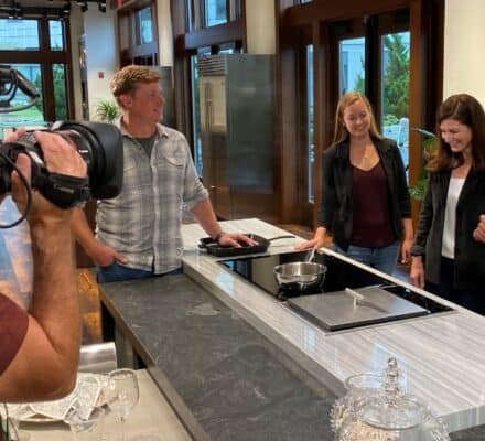 A camera crew films four people gathered around a modern kitchen island, as one man demonstrates cooking on the stovetop using a Wolf appliance.
