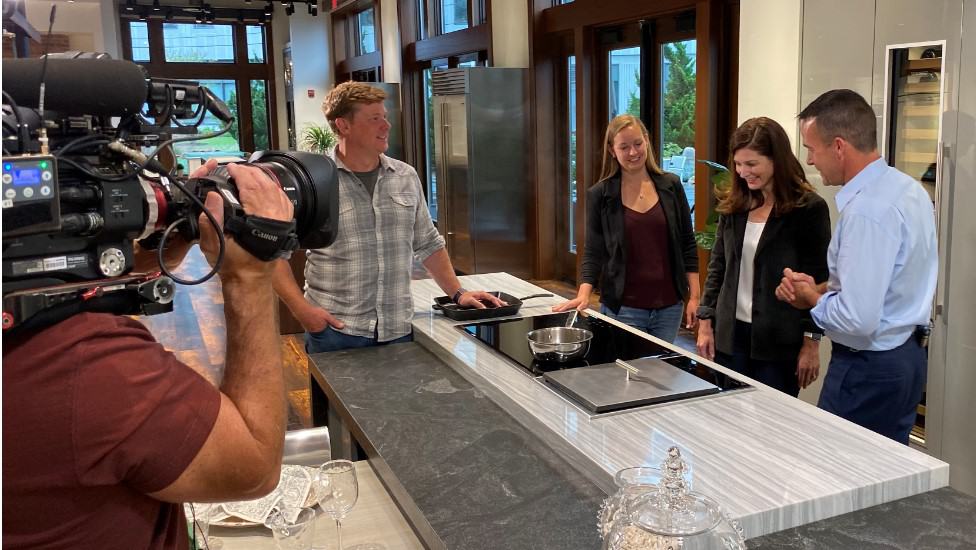 A camera crew films four people gathered around a modern kitchen island, as one man demonstrates cooking on the stovetop using a Wolf appliance.