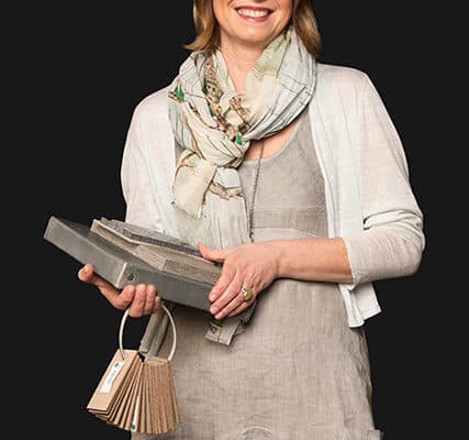 Woman in a light dress and scarf holding tile samples and a material swatch fan, as if selecting finishes for a luxury kitchen appliance in an upscale appliance showroom, all set against a plain black background.