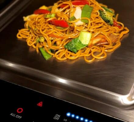 A serving of vegetable lo mein sits directly on a luxury kitchen appliance’s stainless steel stovetop next to a control panel with illuminated buttons.