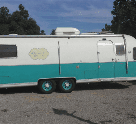 A vintage turquoise and white travel trailer hitched to a white pickup truck, parked on gravel with grass and trees in the background—its cozy interior reminiscent of a kitchen showroom featuring stylish Wolf appliances.