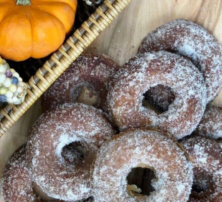 Sugar-coated donuts are arranged on a wooden surface beside a wicker basket with a small pumpkin and Indian corn, creating a scene reminiscent of an autumn display in an upscale appliance showroom.