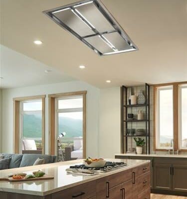 Modern kitchen showroom featuring a large island, built-in stovetop, luxury kitchen appliance, wooden cabinets, open shelving, and large windows with a mountain view. Ceiling-mounted range hood is visible.