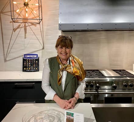 A woman stands and smiles at a kitchen island in a modern kitchen showroom, with a Wolf appliance, vent hood, and decorative light fixture enhancing the luxurious feel in the background.