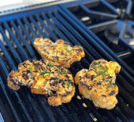 Three seasoned cauliflower steaks are grilling on a stovetop grill, their perfect grill marks and sprinkled herbs highlighted in a modern kitchen showroom featuring gleaming Wolf appliances.