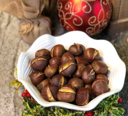 A white bowl filled with roasted chestnuts sits on a festive holly and berry arrangement, with Christmas ornaments in the background—perfect inspiration for your kitchen showroom or luxury kitchen appliance display.