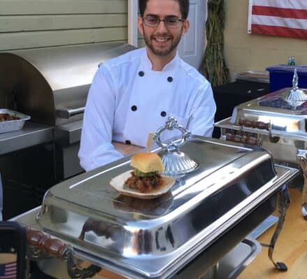 A chef in a white uniform stands behind a serving station with a plated sandwich in front of him, set against the backdrop of an American flag and an appliance showroom featuring Wolf appliances.