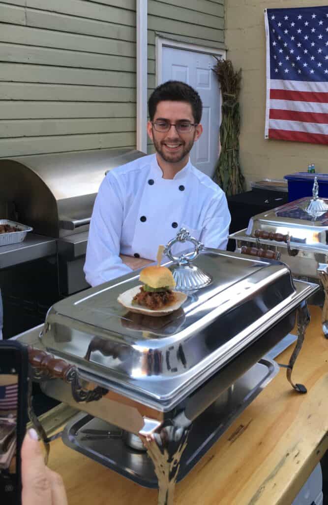 A chef in a white uniform stands behind a serving station with a plated sandwich in front of him, set against the backdrop of an American flag and an appliance showroom featuring Wolf appliances.