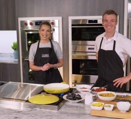 Two people wearing aprons stand in a modern kitchen, smiling at the camera, with cooking ingredients and utensils arranged on the counter in front of a gleaming Sub-Zero appliance.