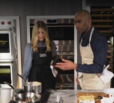 Two people wearing aprons cook together in a modern kitchen, surrounded by sleek luxury kitchen appliances, with pots on the stove and fresh ingredients spread across the counter.