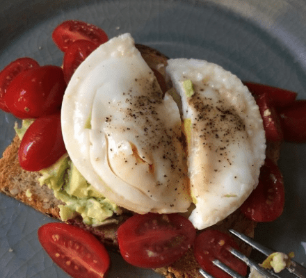 A slice of toast topped with avocado, cherry tomatoes, and a poached egg sprinkled with black pepper on a gray plate—an ideal dish to try after exploring the latest wolf appliance in your local kitchen showroom.
