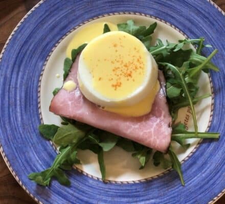 A slice of ham topped with a poached egg and hollandaise sauce sits on a bed of arugula, served on a blue and white plate—perfectly prepared using a Wolf appliance found in any luxury kitchen appliance showroom.