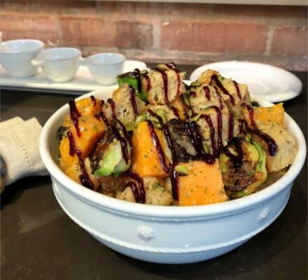 A bowl of salad with chunks of bread, roasted sweet potatoes, brussels sprouts, and a dark balsamic glaze on top sits on a dark countertop, ready to enjoy fresh from your luxury kitchen appliance.
