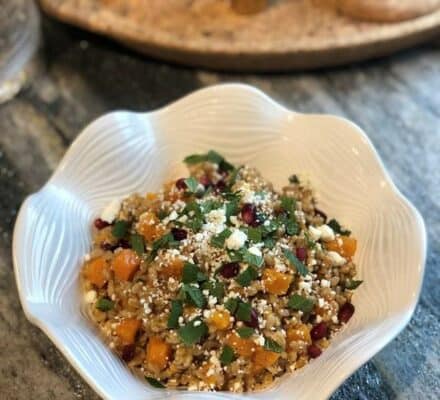 A white bowl filled with a grain salad featuring chopped vegetables, herbs, feta cheese crumbles, and pomegranate seeds is placed on a gray countertop in a luxury kitchen appliance showroom.
