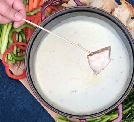 A person dips a piece of bread into a pot of cheese fondue, surrounded by assorted vegetables and bread on a wooden board in a stylish kitchen showroom.