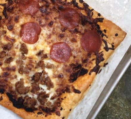 A rectangular pizza with melted cheese, pepperoni slices, and sausage pieces bakes to perfection on a sheet using a Wolf appliance—essential for any luxury kitchen.