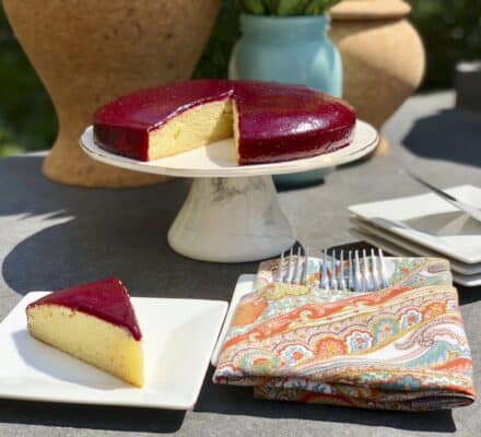 A round cake with a glossy berry topping sits on a cake stand in a luxury kitchen appliance showroom; a single slice is served on a plate next to forks and a patterned napkin.