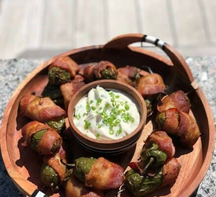 A wooden tray from a kitchen showroom displays bacon-wrapped jalapeño poppers arranged around a bowl of dip topped with chopped chives.