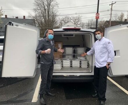 Two men wearing masks and gloves stand beside an open van filled with stacked food containers and boxes in a parking lot outside an appliance showroom.