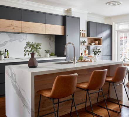 Modern kitchen with a marble island, luxury kitchen appliances, three brown leather stools, black cabinets, wood accents, and potted plants, with natural light coming through large windows.