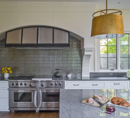 Modern kitchen with stainless steel and sub zero appliances, a gray tile backsplash, granite countertops, and a gold pendant light. Fresh pastries and flowers are on the island near a window.