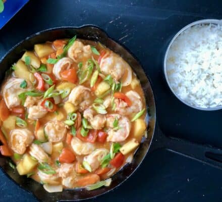 A skillet filled with shrimp, pineapple, and vegetables in sauce sits next to a small pot of cooked white rice on a dark surface, as seen in a luxury kitchen showroom featuring Sub-Zero and Wolf appliances.