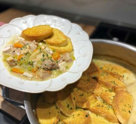 A white plate holds a serving of chicken stew with vegetables and toasted bread rounds, next to a pan filled with more stew topped with toasted bread slices—an inviting scene perfect for any kitchen showroom display.