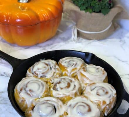 Seven iced cinnamon rolls sit in a black cast iron skillet on a marble countertop, with a pumpkin-shaped container, a potted plant, and the subtle reflection of an appliance showroom featuring Sub-Zero appliances in the background.