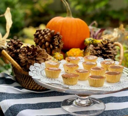A cake stand with mini desserts on a doily is set on a striped cloth, accented by a basket of pinecones, gourds, and a pumpkin—a scene fit for any luxury kitchen appliance display or upscale kitchen showroom.