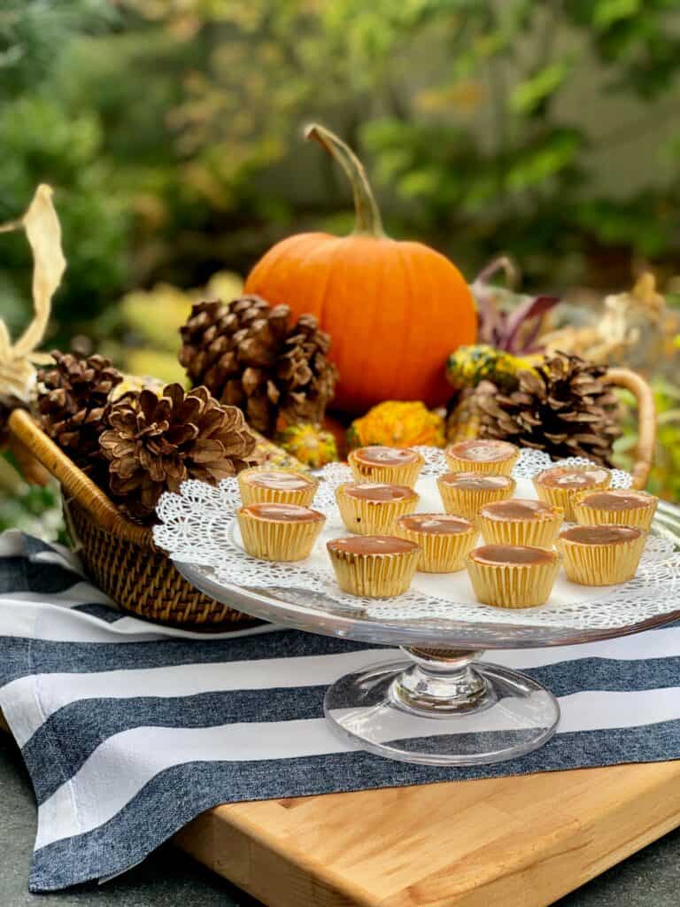 A cake stand with mini desserts on a doily is set on a striped cloth, accented by a basket of pinecones, gourds, and a pumpkin—a scene fit for any luxury kitchen appliance display or upscale kitchen showroom.